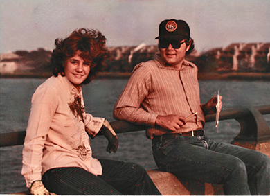 An aged photo of two people sitting outside. David is looking across at Amy who is wearing a soild shirt and gloves and smiling at the camera.